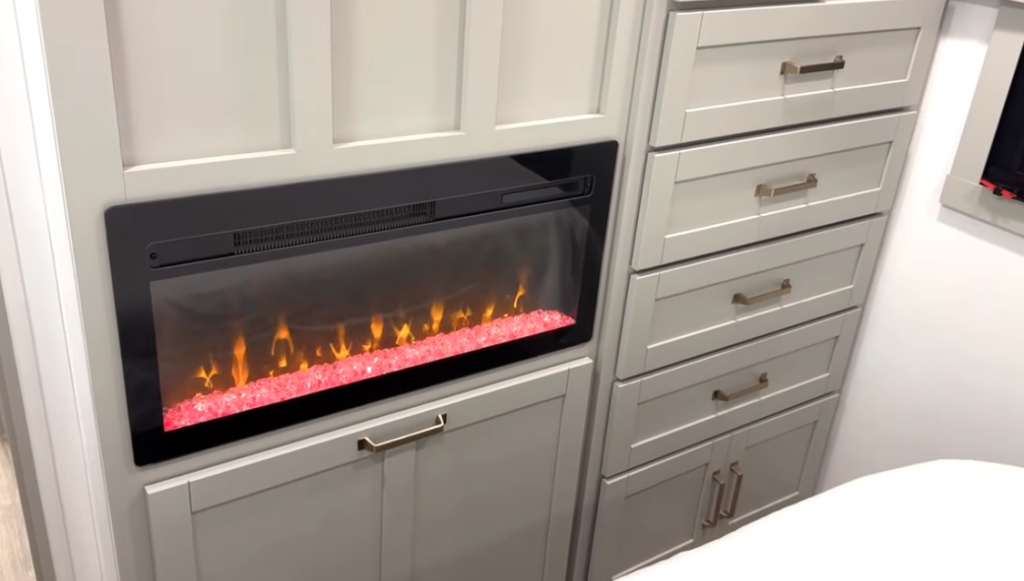 Bedroom storage wall in the 2026 Brinkley Model Z Air featuring a built-in electric fireplace with glowing ember bed, tall dresser drawers, and shaker-style cabinetry for expanded RV organization.