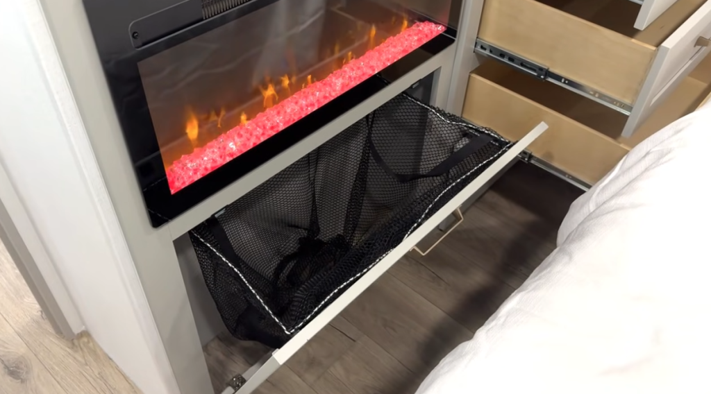 Pull-out RV bedroom laundry hamper in the 2026 Brinkley Model Z Air located beneath the built-in electric fireplace, featuring a mesh basket for easy clothes storage next to deep dresser drawers.