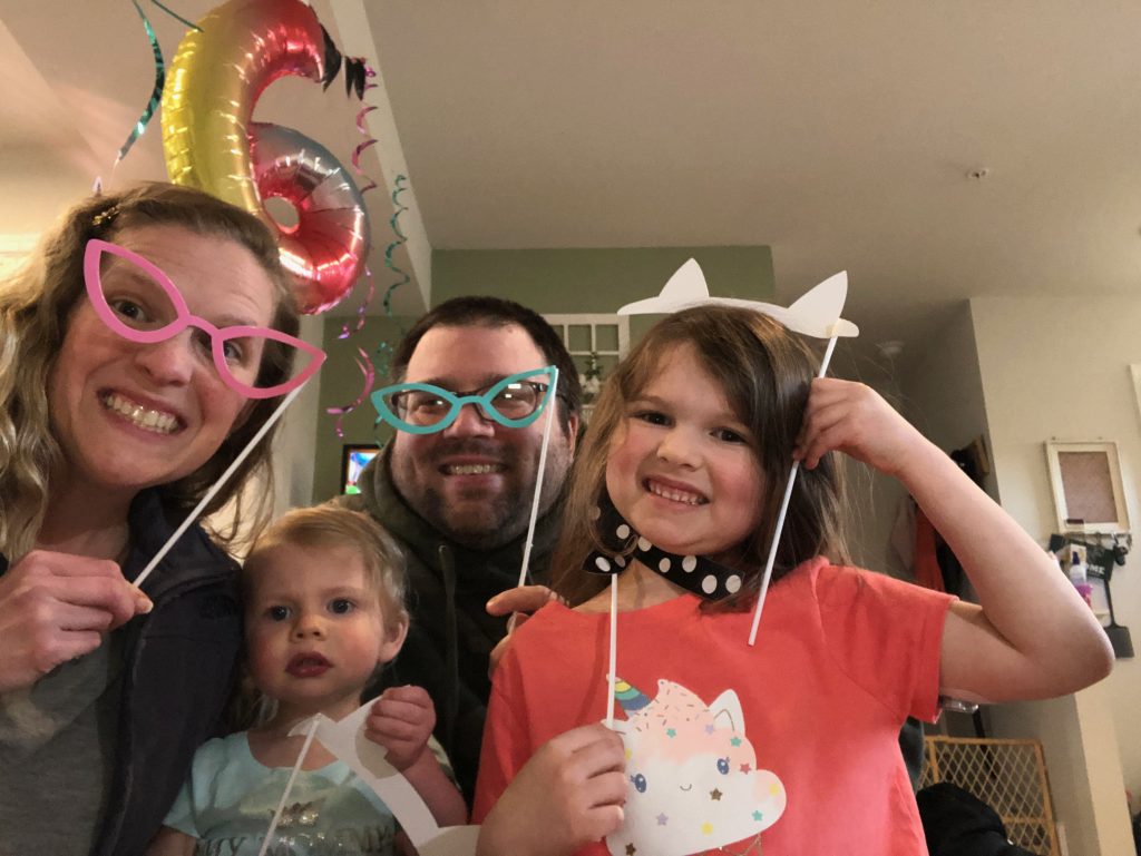 Family celebrating indoors with playful photo-booth props, colorful glasses, and party decorations, smiling together during a fun birthday celebration.