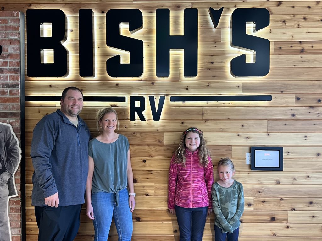 Family standing in front of the illuminated Bish’s RV wood-panel wall, smiling together inside the dealership for a welcoming customer experience photo.