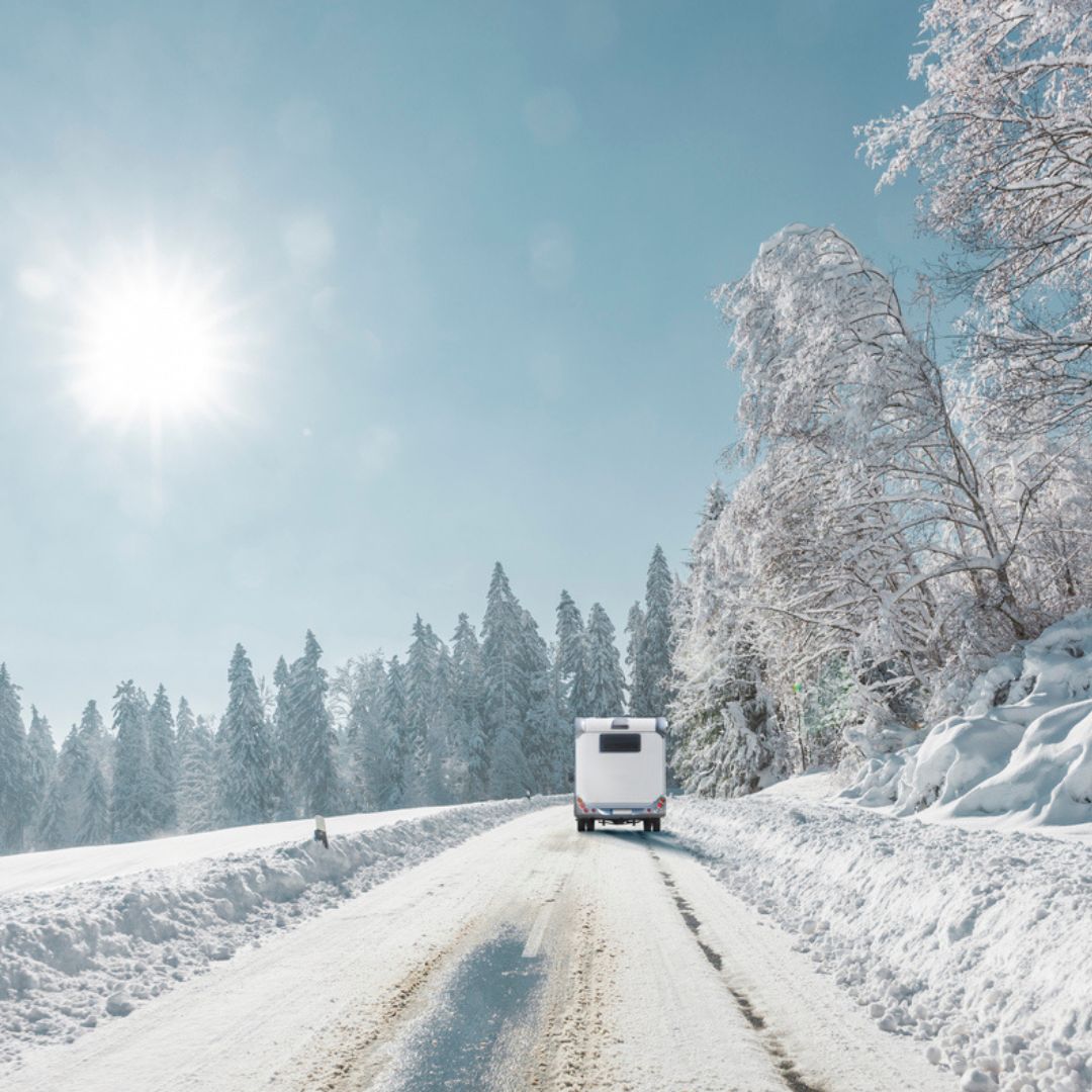 RV driving down a snow-covered forest road on a bright winter day, surrounded by frost-covered trees and clear blue skies — symbolizing winter RV travel and adventure.