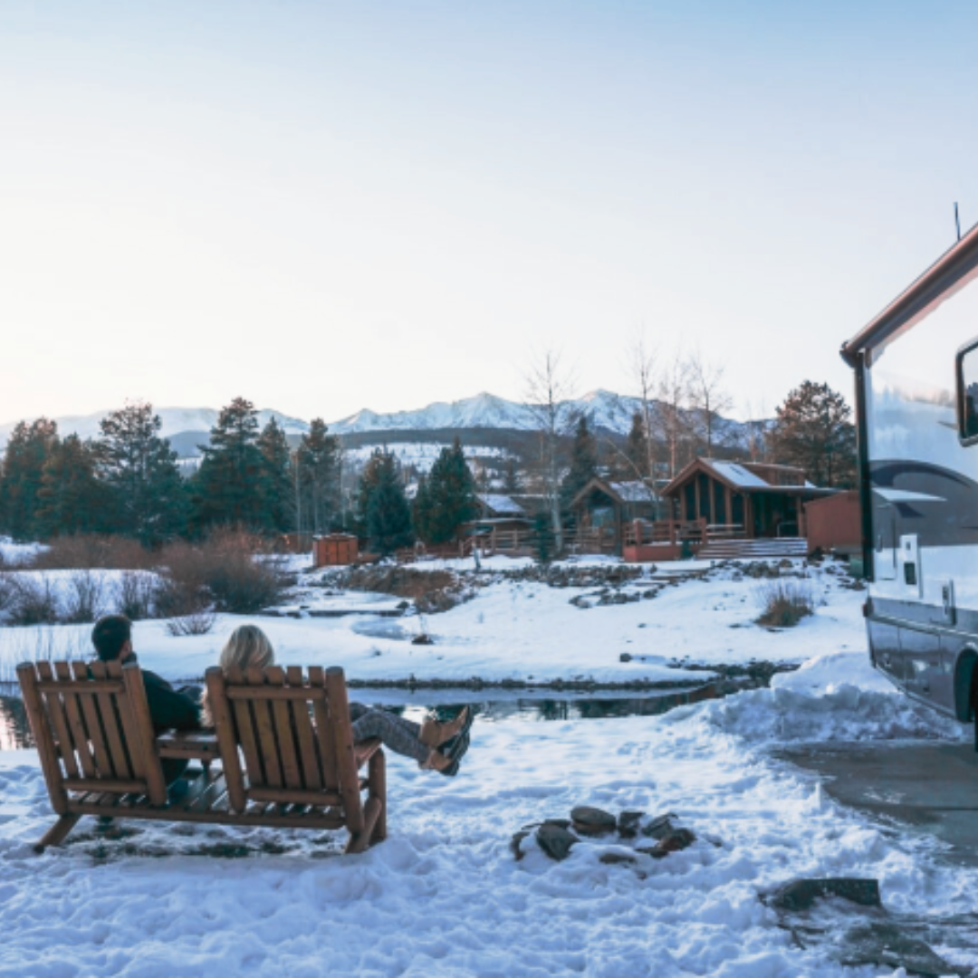 Couple sitting on wooden chairs beside their RV, overlooking a snowy campground with cozy cabins and snow-covered mountains in the distance during winter camping season.