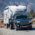 Heavy-duty GMC truck towing a Jayco fifth-wheel RV through a scenic mountain landscape on a rural road.