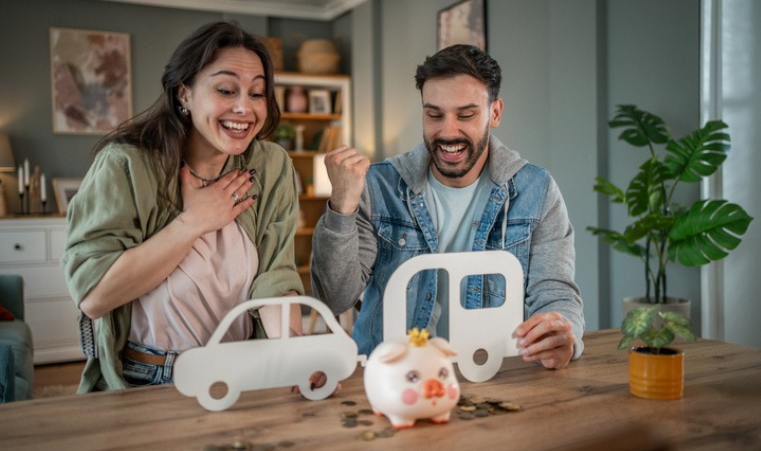 Excited couple sitting at a table celebrating savings, with paper cutouts of a car and RV next to a piggy bank, symbolizing budgeting for travel or RV ownership.