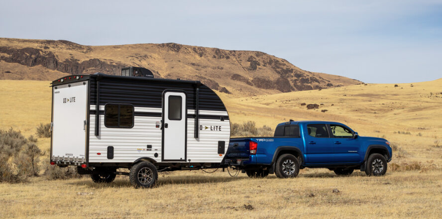 Toyota Tacoma towing a lightweight Wayfinder Go Lite travel trailer through an open desert landscape with rocky hills in the background.