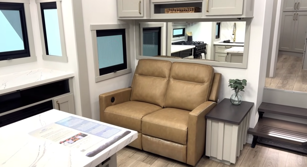 RV living area with a tan dual-reclining loveseat, side table with plant, large wall mirror, and modern cabinetry near the kitchen and entry steps.