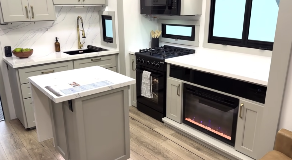 Modern RV kitchen with center island, marble-look countertops, farmhouse sink, gas stove, microwave, and built-in electric fireplace beneath the counter.