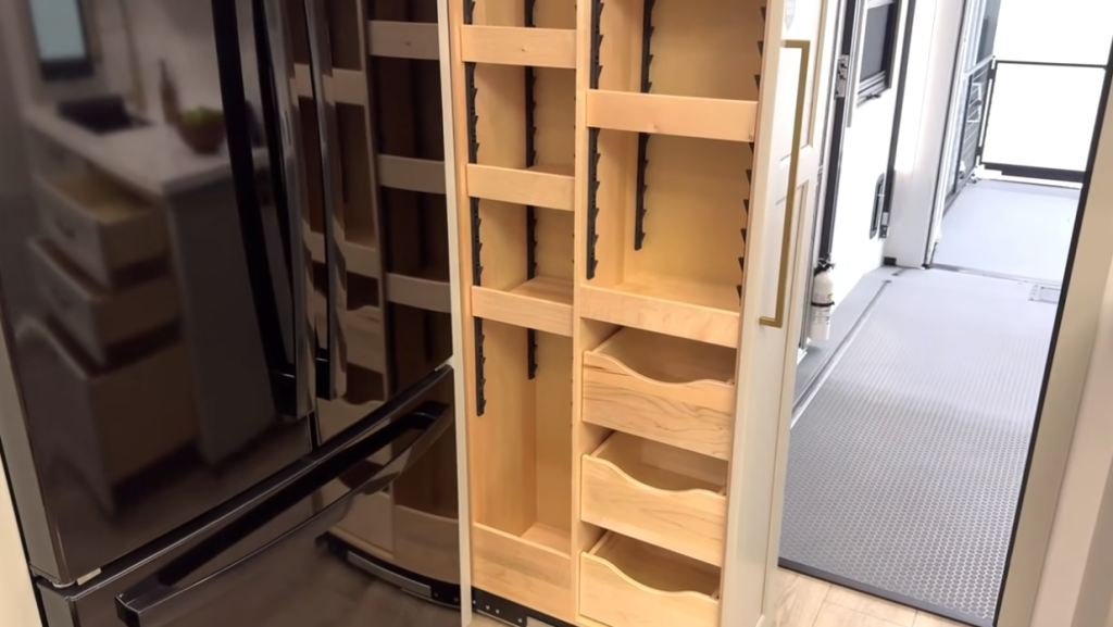 Slide-out pantry beside a residential refrigerator inside a Brinkley RV, featuring adjustable shelves and built-in storage bins for organized food and kitchen supplies.