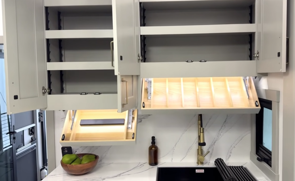 Open overhead cabinets and fold-down drying racks in a modern Brinkley RV kitchen, showcasing added storage, organization, and functional workspace above the sink.