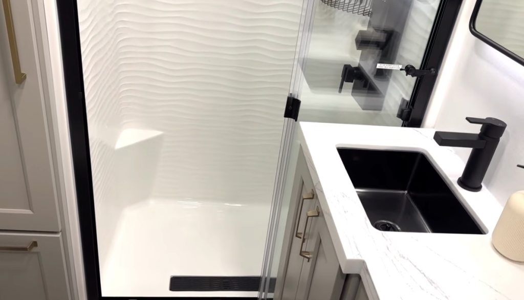 Modern RV bathroom with a spacious white shower featuring textured walls and a built-in seat, next to a sleek vanity with a black sink and matte black faucet.