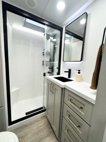 Bright, modern RV bathroom with a glass-enclosed textured shower, skylight, matte black fixtures, spacious vanity storage, and light wood-look flooring.