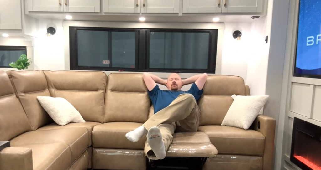 Josh the RV nerd on the reclining L-shaped sofa in the Brinkley Model Z 3200 fifth wheel, showing the spacious RV living area with overhead cabinets, large windows, and entertainment center.