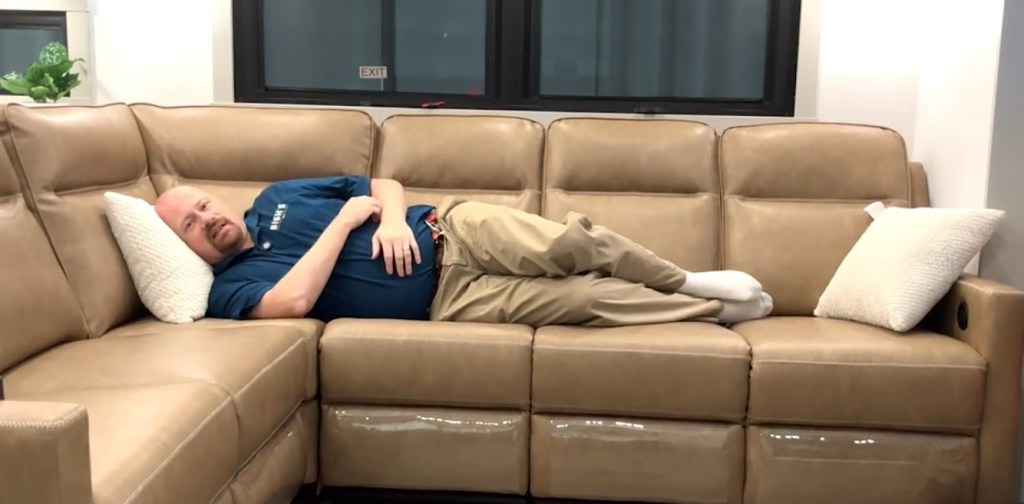 Josh the RV nerd lying on the large tan L-shaped reclining sofa in the Brinkley Model Z 3200 fifth wheel, demonstrating the spacious RV living room seating