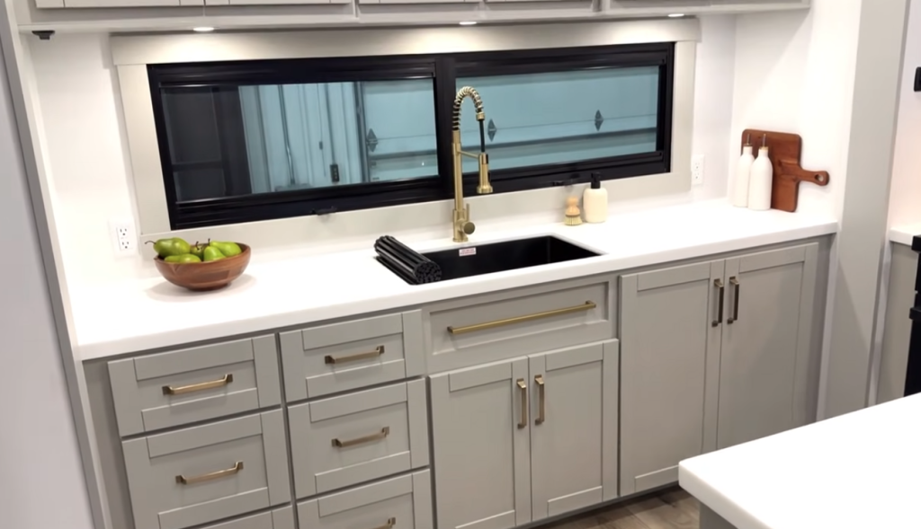 Brinkley Model Z 3200 kitchen featuring solid-surface countertops, black farmhouse sink with gold pull-down faucet, spacious gray cabinetry, and large horizontal window for natural light.