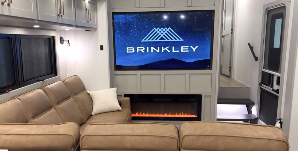 Brinkley Model Z 3200 RV living room with large entertainment center, LED fireplace, big-screen TV, overhead cabinets, and tan L-shaped reclining sofa