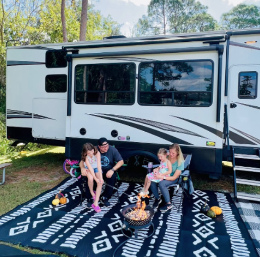 Family sitting around a small campfire on an outdoor rug beside their travel trailer at a wooded campsite, enjoying RV camping together