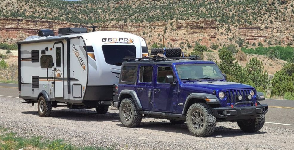 Jeep Wrangler towing a Rockwood Geo Pro travel trailer on a scenic desert highway with red rock cliffs in the background.