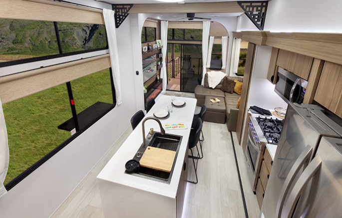 Modern travel trailer interior featuring a center island kitchen with residential appliances, large panoramic windows, cozy living area with sofa, and rear patio deck