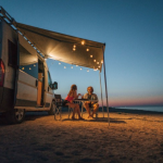 Couple relaxing under an awning with string lights beside their camper van while beach camping at sunset.