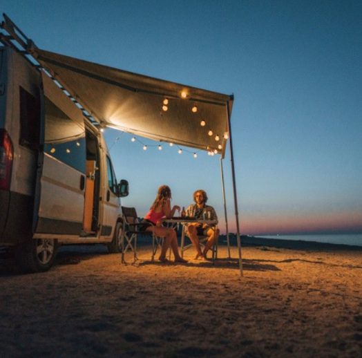 Couple relaxing under an awning with string lights beside their camper van while beach camping at sunset.