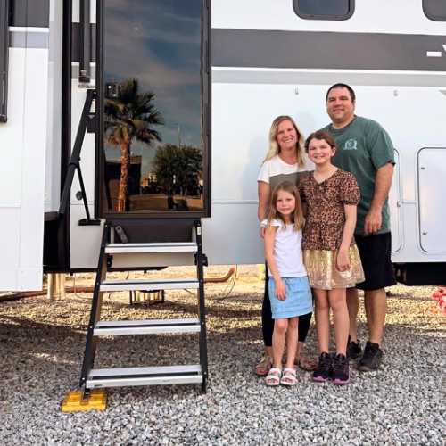 Family of four standing in front of their fifth wheel RV with entry steps visible, smiling at a campground site, representing family RV travel, ownership, and outdoor adventures.