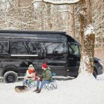 Couple sitting beside a black camper van in the snow with a dog, enjoying a winter RV camping trip in a forest setting.