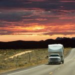 Pickup truck towing a fifth wheel RV down an open highway at sunset with desert landscape and dramatic colorful sky in the background.