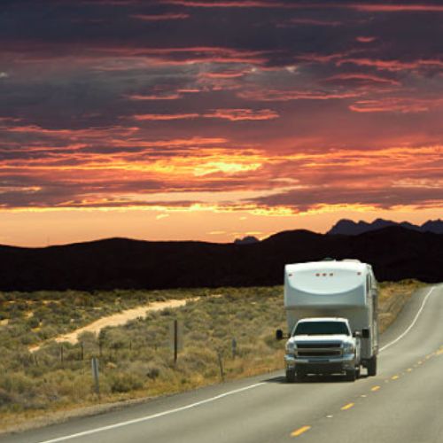 Pickup truck towing a fifth wheel RV down an open highway at sunset with desert landscape and dramatic colorful sky in the background.