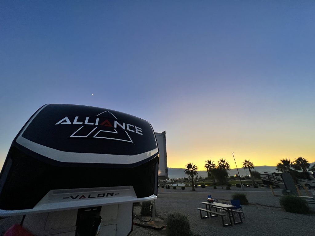 Sun going down just behind the mountains while showing the moon sitting above the front cap of an Alliance Valor toyhauler.