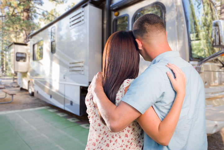 Couple standing arm in arm while looking at their parked fifth wheel RV at a wooded campground, symbolizing RV ownership and outdoor travel lifestyle.
