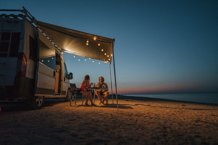 Couple relaxing under an awning with string lights beside their camper van while beach camping at sunset.