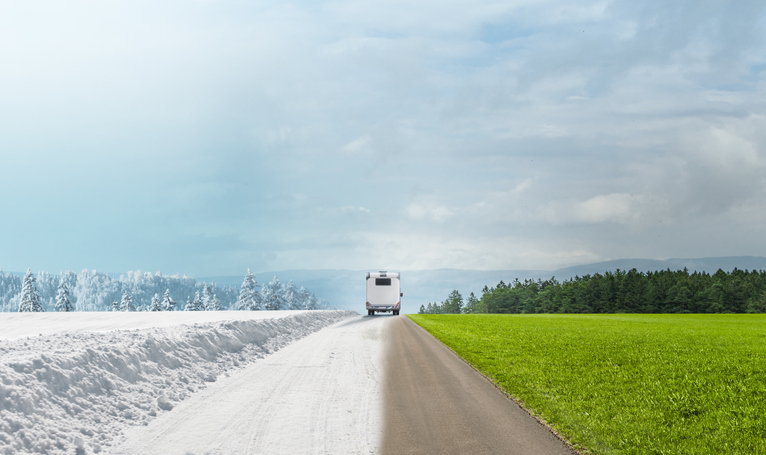 RV driving down a road transitioning from winter snow to green summer landscape, symbolizing year-round RV travel and seasonal camping