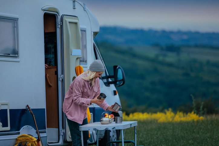 Woman cooking at an outdoor table beside her RV while camping in a scenic hillside meadow during sunset.