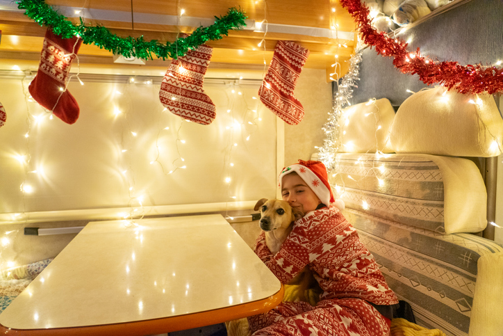 Child in Christmas pajamas hugging a dog inside a festively decorated RV dinette with stockings, garland, and holiday lights