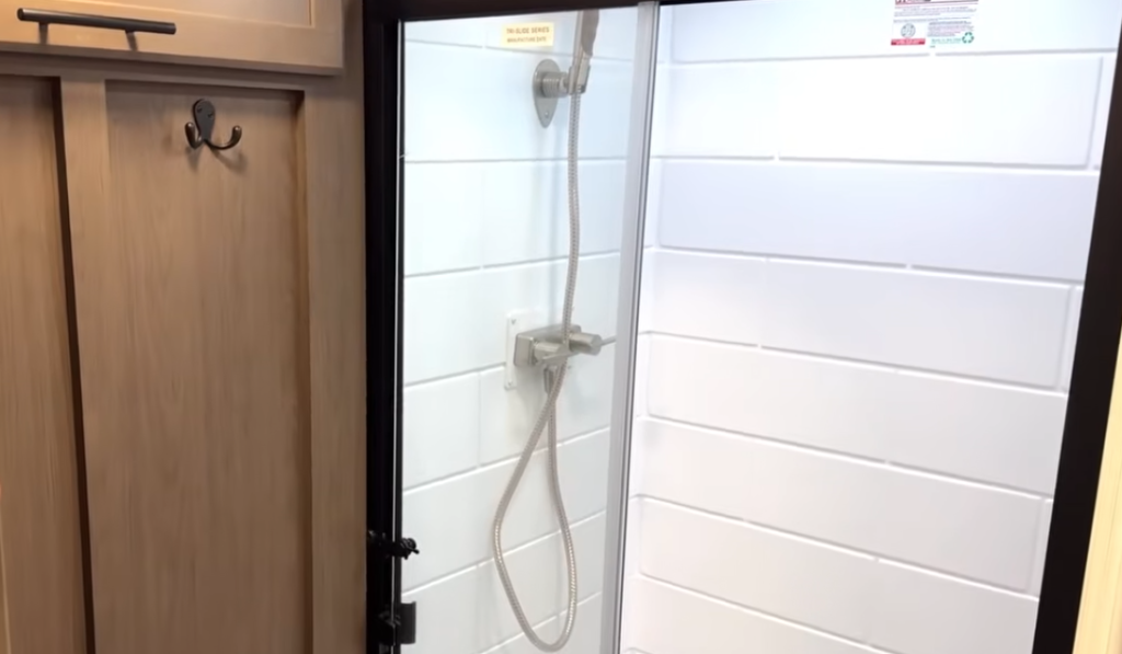 RV bathroom shower with glass door, white subway-style wall panels, adjustable handheld showerhead, and nearby storage hooks, designed for easy cleaning and a modern, residential-style bathing experience in an RV.