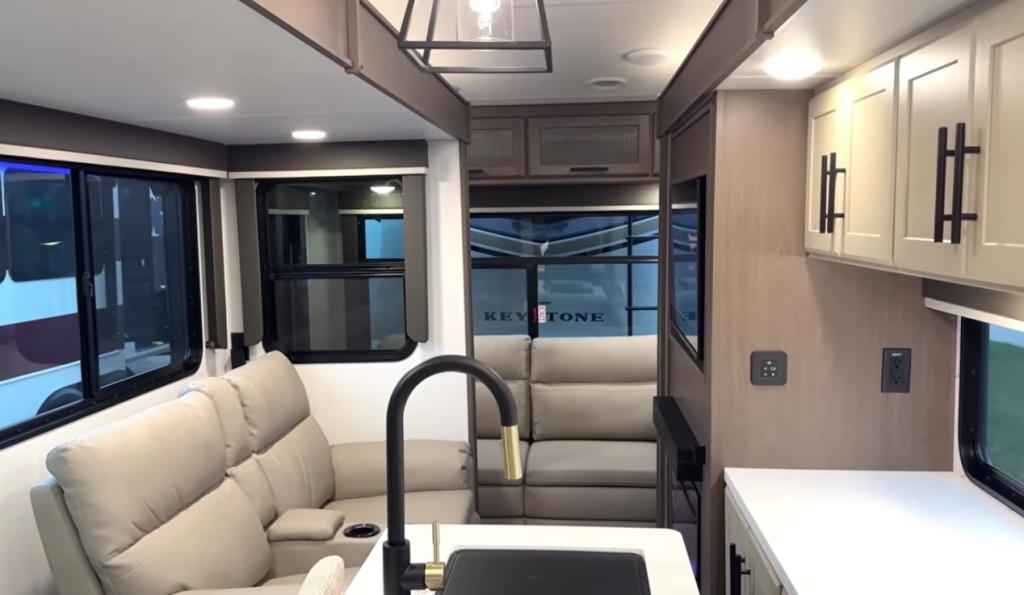 Keystone Cougar fifth wheel interior showing modern living room and kitchen layout with reclining sofa, large windows, solid-surface countertops, and contemporary cabinetry designed for half-ton towable RVs.