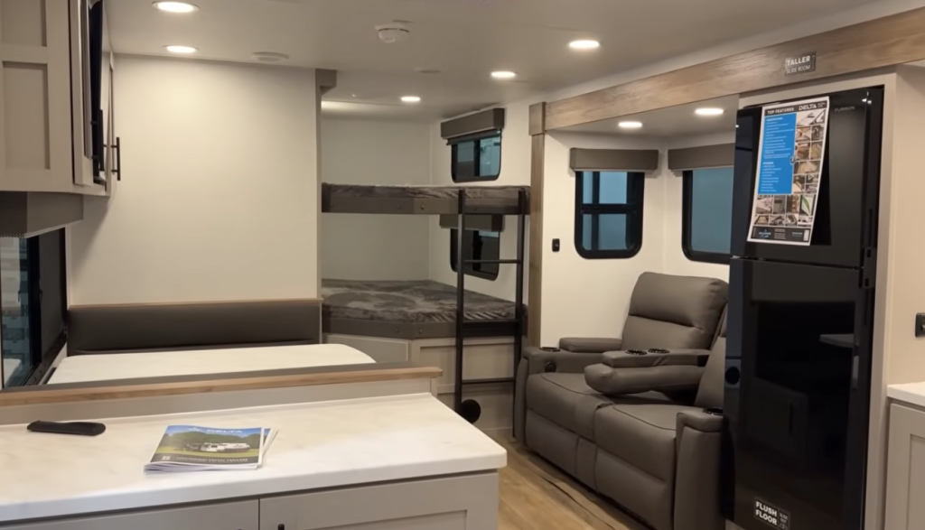 Open-concept travel trailer interior featuring rear double bunk beds, a reclining theater-style sofa, full kitchen with island countertop, overhead LED lighting, and modern cabinetry in a lightweight RV floorplan.