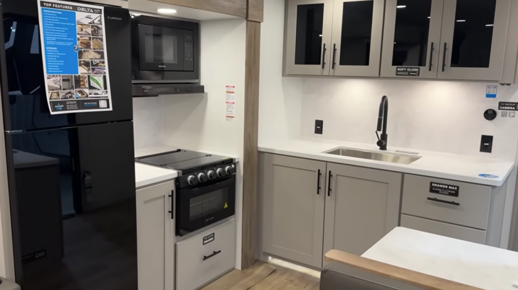 Modern RV kitchen with full-size refrigerator, propane oven and stovetop, microwave, deep stainless sink with matte black faucet, soft-close cabinets, and solid-surface countertops in a contemporary travel trailer.