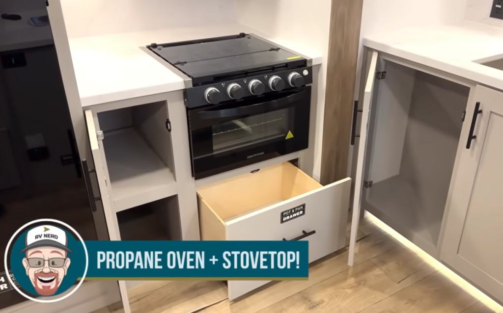 RV kitchen propane oven and three-burner stovetop with glass cooktop cover, pull-out pots and pans drawer, soft-close cabinets, and solid-surface countertops in a modern travel trailer kitchen.