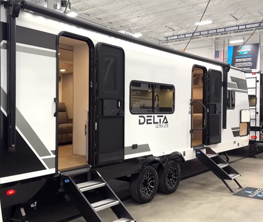Alliance Delta Ultra Lite travel trailer exterior with dual entry doors, aluminum steps, tandem axle wheels, large awning, and modern black-and-white graphics displayed indoors at an RV showroom.