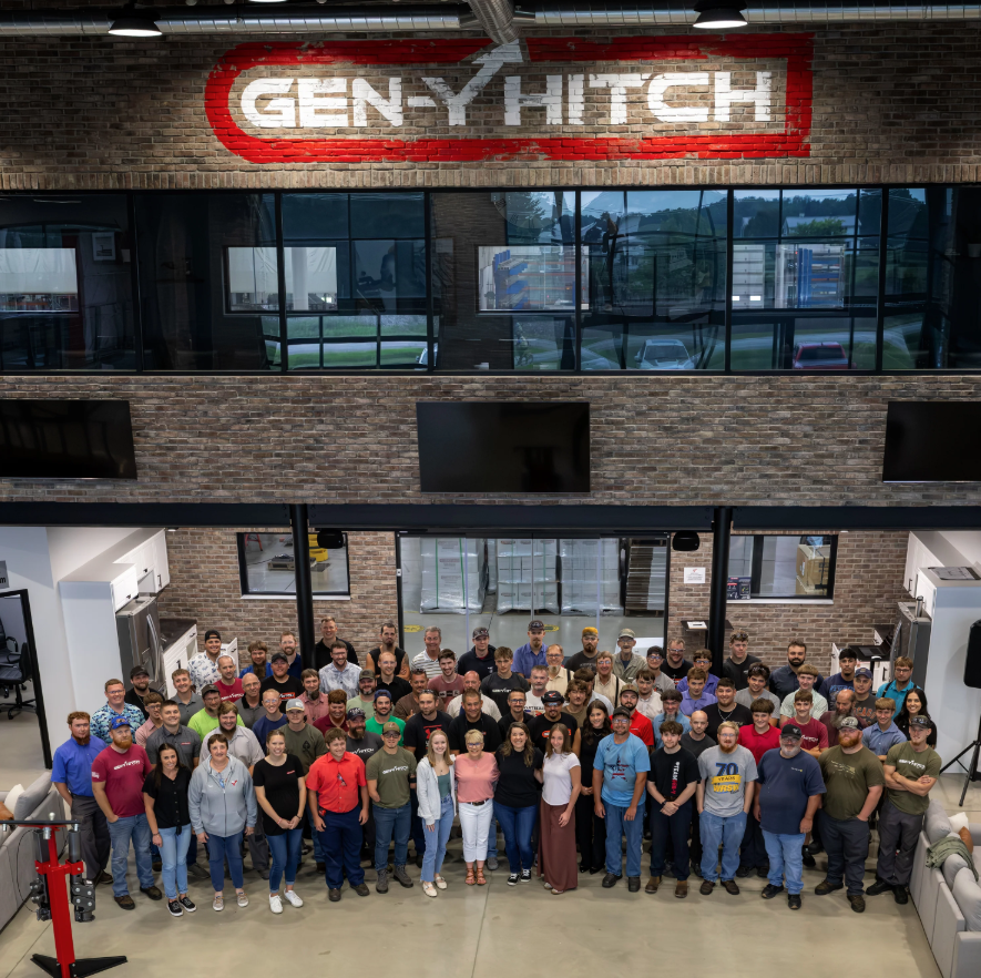 Gen-Y Hitch team standing inside the company’s manufacturing facility with large Gen-Y Hitch logo on the wall