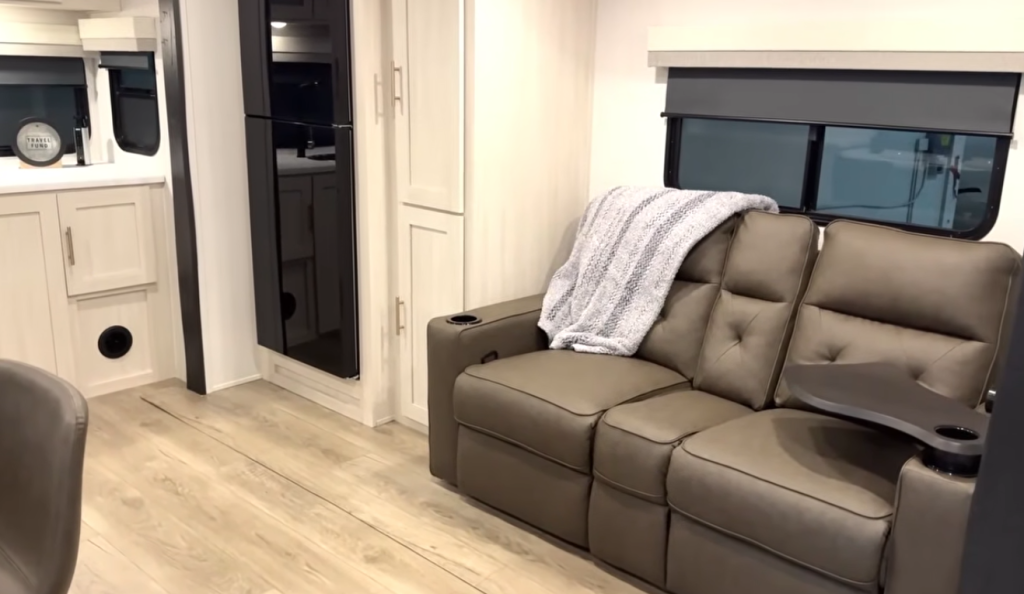 Modern RV living area with theater seating sofa, refrigerator, storage cabinets, and light wood-look flooring