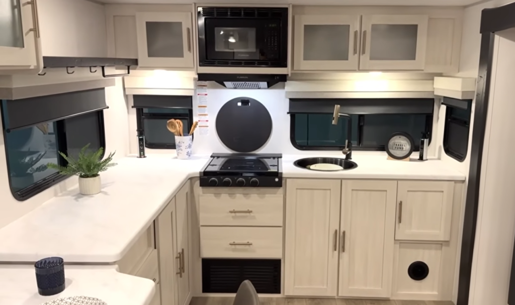 Modern RV kitchen with three-burner stove, microwave, round sink, and light-colored cabinets with solid-surface countertops
