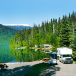 RV campsite beside a scenic mountain lake with trees, picnic table, and camper parked near the water