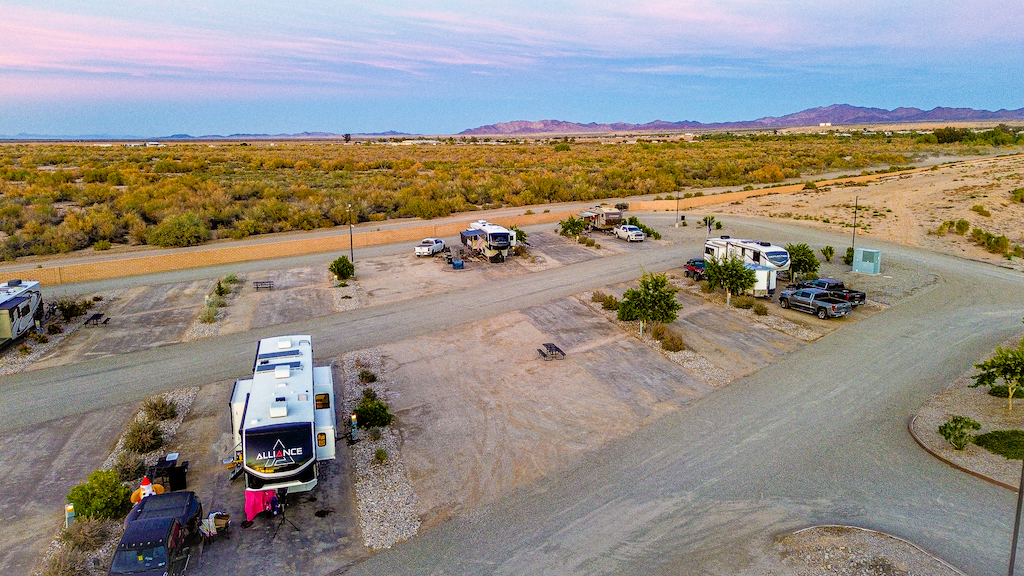 Aerial view of RV campground with multiple trailers and vehicles parked in a desert landscape at sunset.