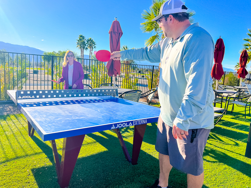 Guests playing outdoor ping pong at an RV resort with palm trees, patio seating, and mountain views
