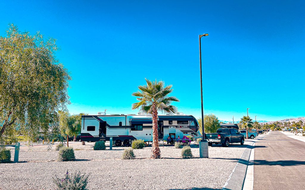 Fifth wheel RV parked at a sunny desert RV park with palm trees, wide roads, and full hookup sites