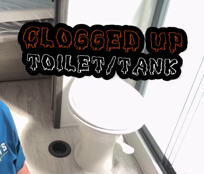 Graphic reading “Clogged Up Toilet/Tank” in bold orange and white text over an RV bathroom scene, showing a compact RV toilet and floor drain, illustrating black tank or toilet clog issues.