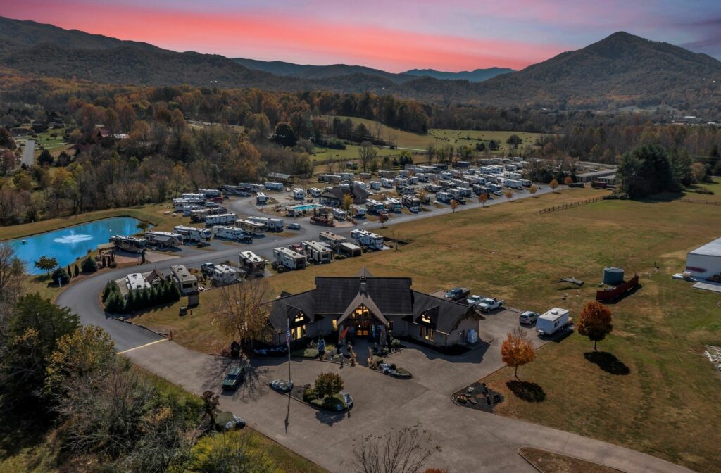 Aerial view of a scenic mountain RV resort at sunset, featuring a central lodge, full-hookup campsites, a small pond, and rolling hills of the Blue Ridge Mountains in the background.