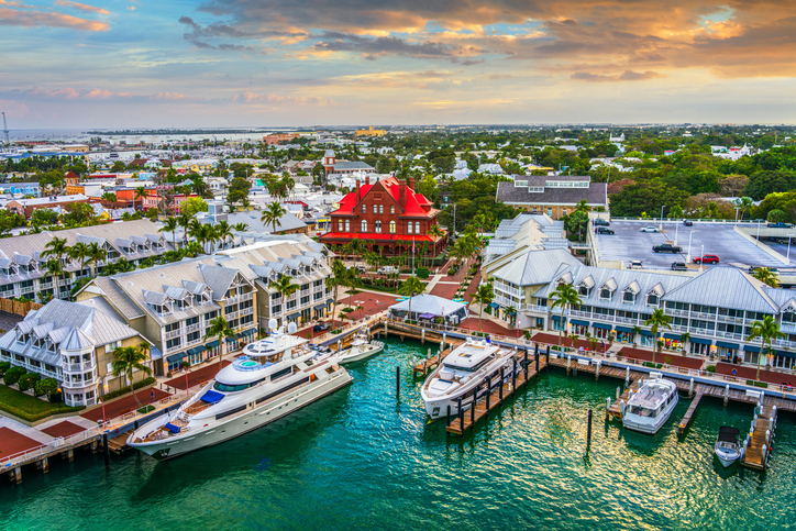 Aerial view of historic Key West waterfront at sunset, featuring the red-roofed Key West Aquarium, marina docks with luxury yachts, palm-lined streets, and colorful island architecture along the harbor.